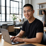 A smiling man with glasses and a black t-shirt sits at a wooden desk in a bright office, typing on a silver laptop.